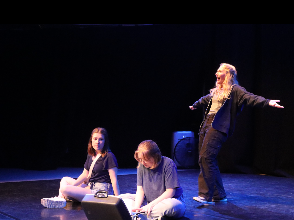 Three young women performing on a dark stage. One woman is standing with arms outstretched and mouth open, appearing expressive. Two women are sitting on the floor, one looking at the camera, and the other looking down at an electronic device.