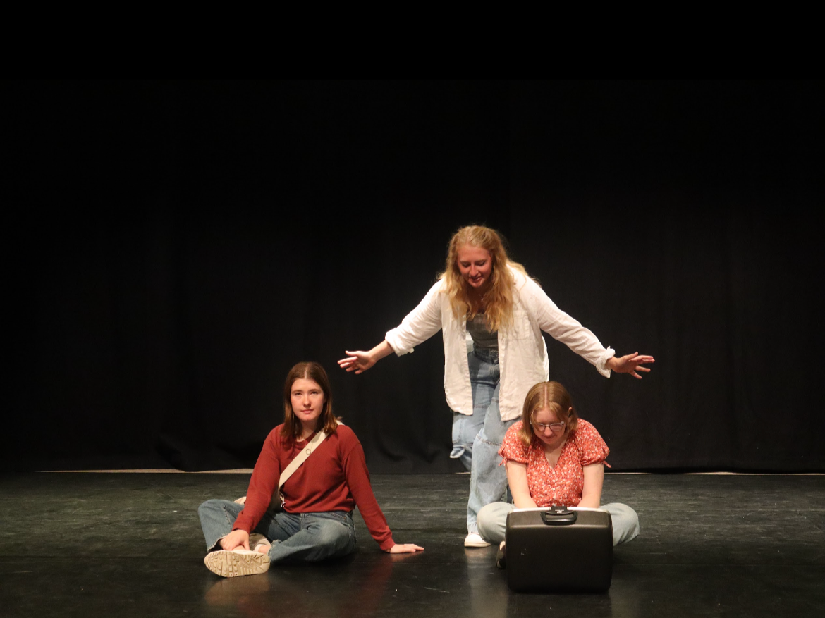 Three young women on a stage with a black background, two sitting on the floor and one standing behind them with arms outstretched.