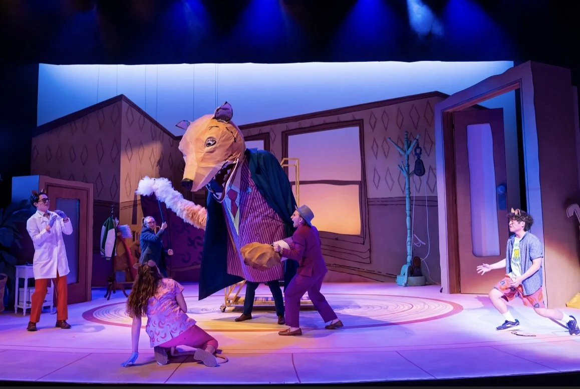 A theatrical stage with a large animal character costume, resembling a fox, in the center. Several people in colorful costumes are interacting with the character, with some kneeling or stretching. The background features cartoonish house set pieces, a window with sunset scenery, and a coat rack. The scene is lit with vibrant, colorful stage lighting.