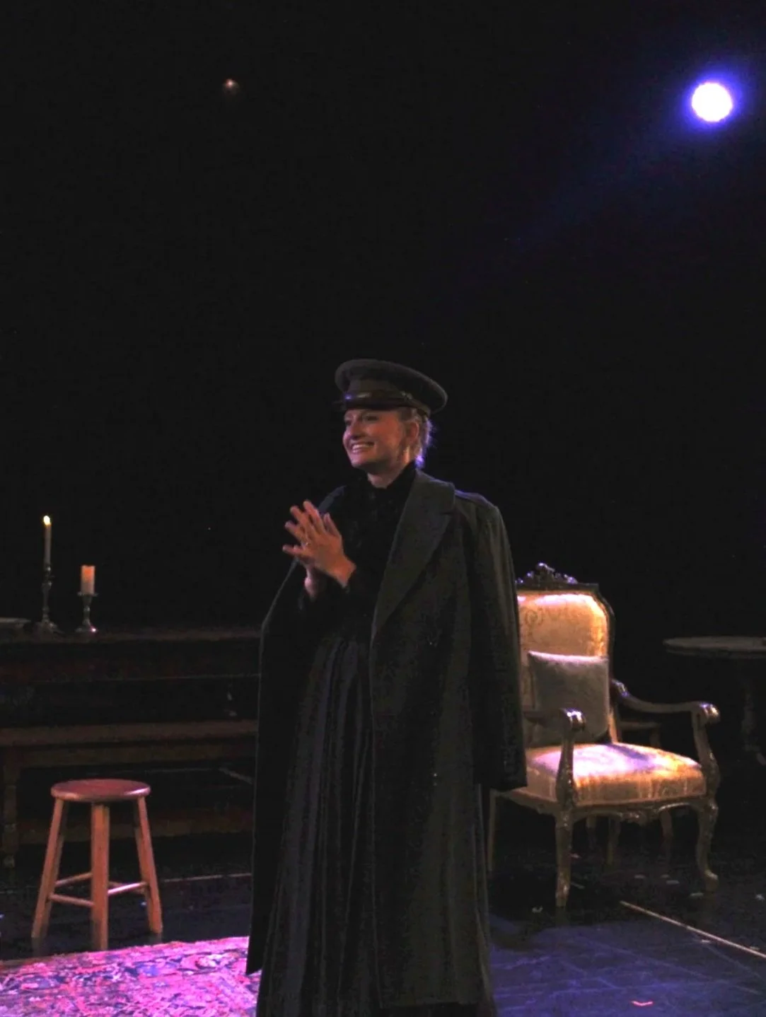 A woman wearing a dark graduation gown and cap standing on a stage, smiling with hands clasped, with an audience area dimly lit and a full moon visible in the night sky.