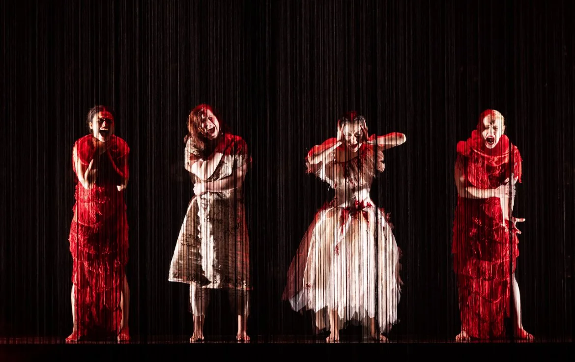 Four women performing on stage during a rainstorm, dressed in vintage-style and brightly colored costumes, with dark background.