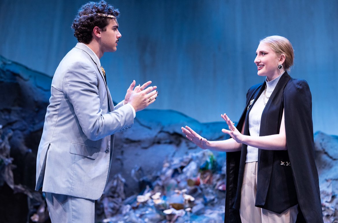 A man and a woman are having a conversation on stage with a backdrop that appears to be a polluted environment or a landfill, with trash and debris visible.