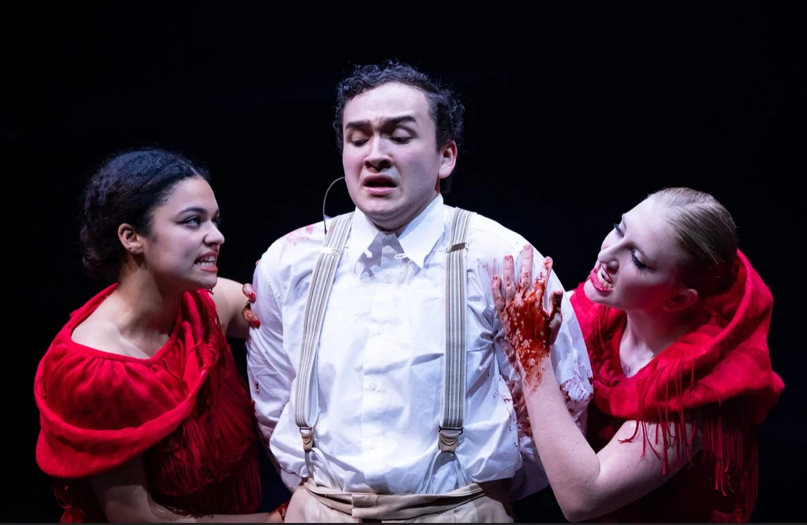 Three actors on stage, two women and one man, in a theatrical scene with intense expressions. The man appears distressed, with blood on his shirt and hand, while the women are holding his shoulders and are also emotional.