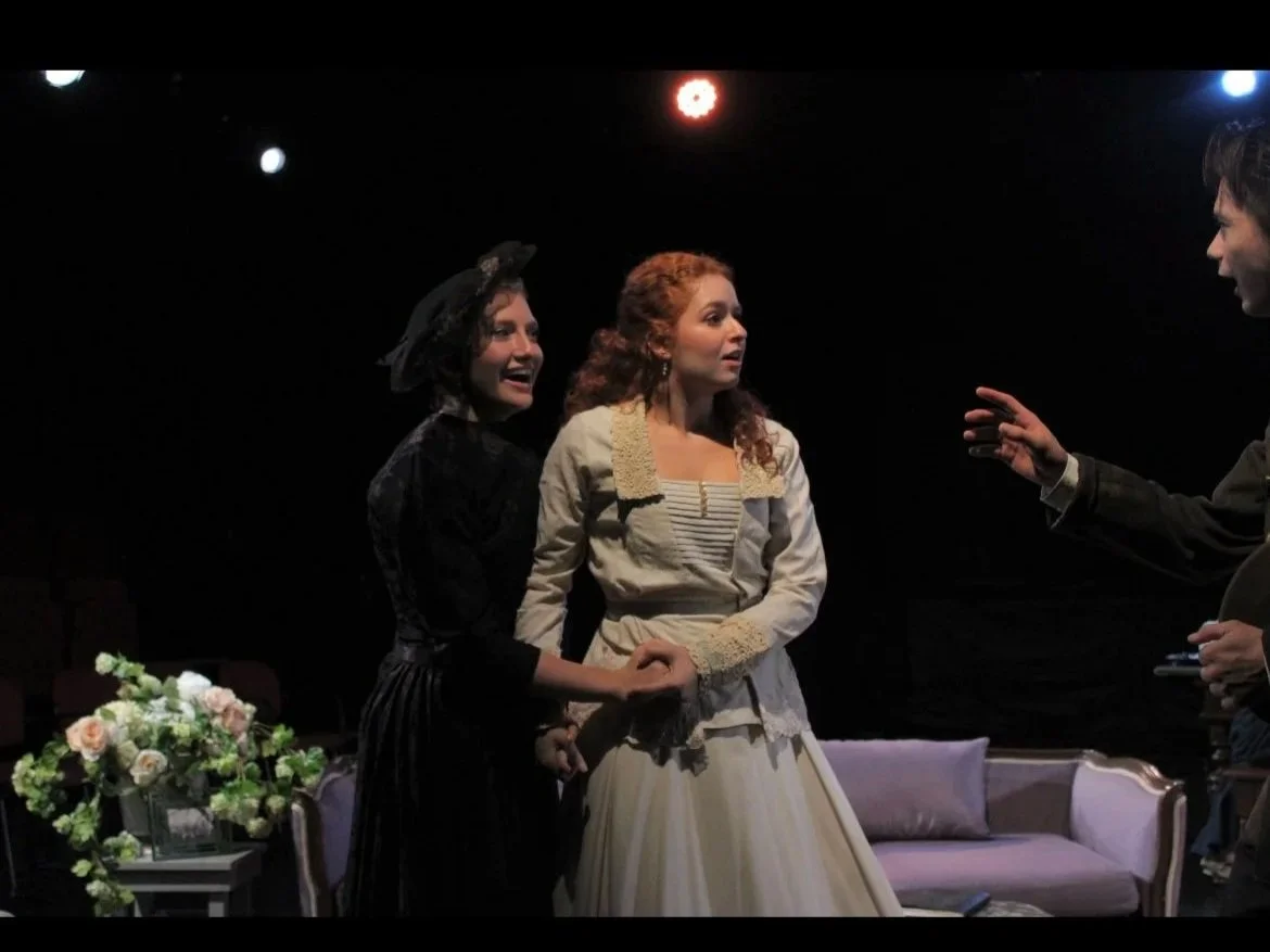 Three actors on stage during a theatrical performance, with two women and a man. The women are holding hands, one wearing a black dress with a crown and the other in a light-colored dress. The man is gesturing toward them.