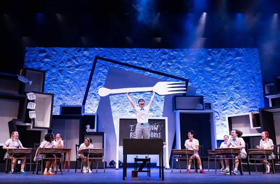 A theatrical performance with children in school uniforms on stage, a woman standing on a desk with her arms raised, a large spoon and fork graphic in the background, and a set designed to resemble a classroom.