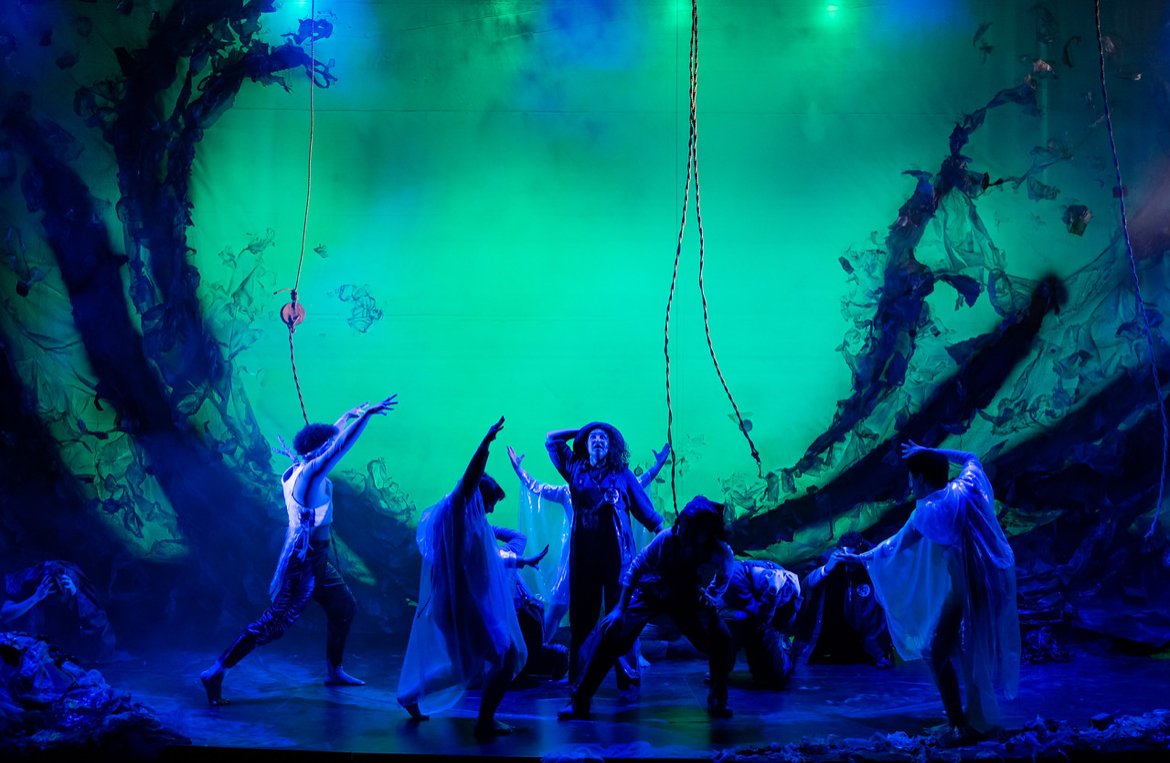 Theater performance with actors on stage in dark costumes, some wearing flowing white garments, under vibrant green and blue lighting, with a painted backdrop resembling a forest.