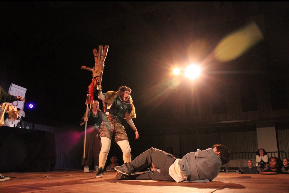 People are performing a theatrical scene on stage, with one person lying on the floor and another person standing over them, expressing a dramatic gesture. Others are in the background holding props, including a sign.