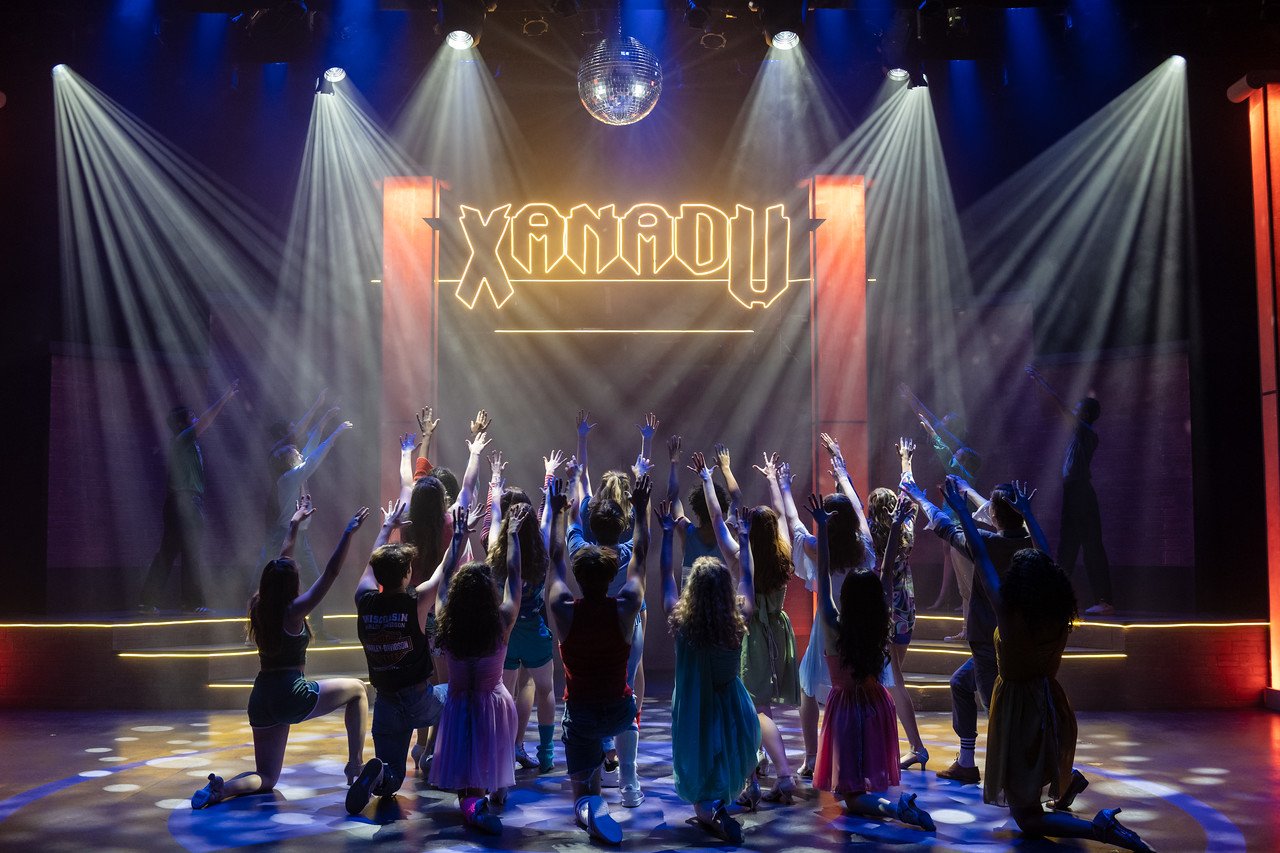 Group of children on stage performing dance in front of a neon sign that reads "XANADU" with stage lights and a disco ball above.
