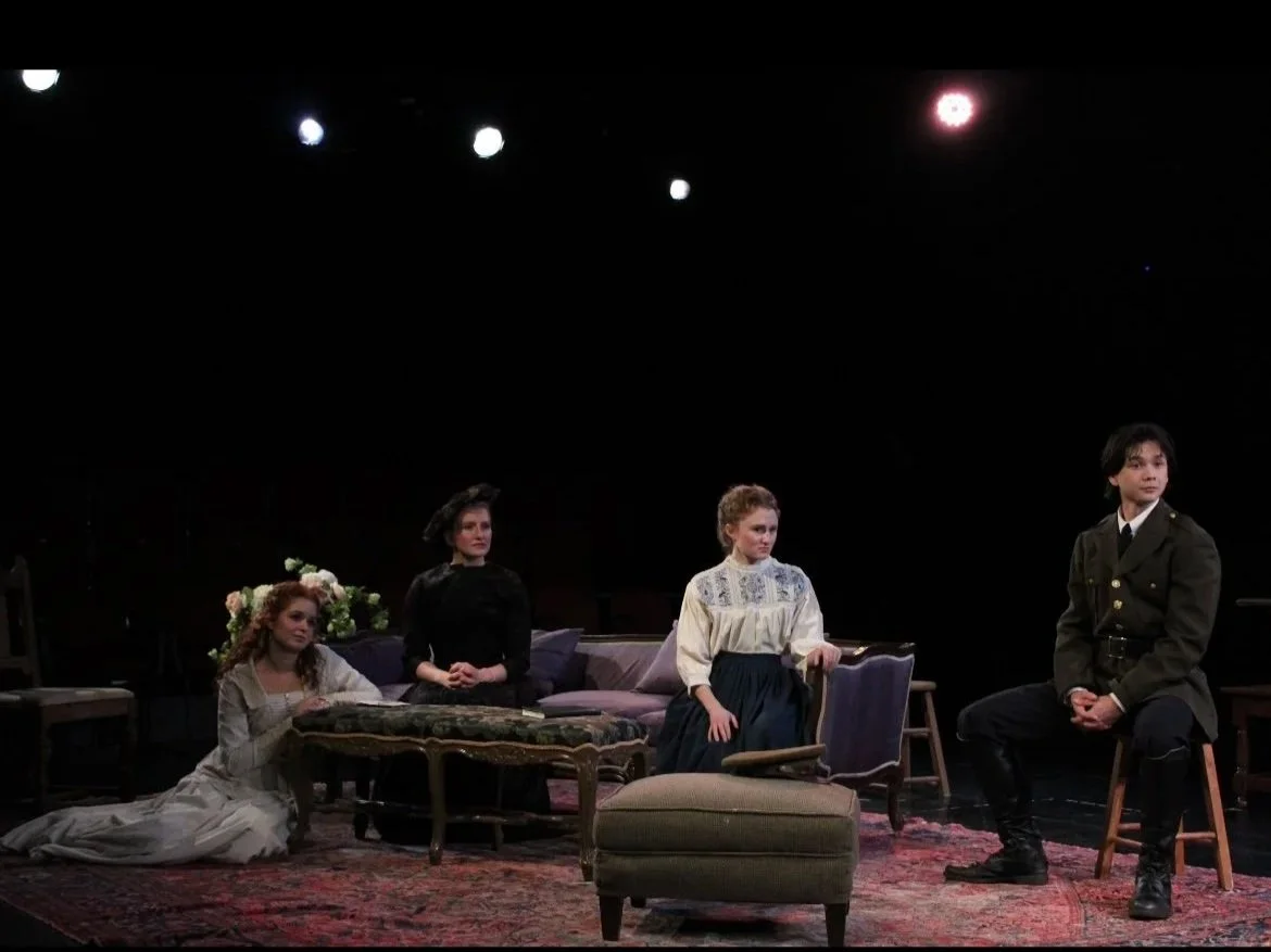 Four actors on stage in period costumes, sitting and standing around a vintage coffee table, with a dark background and stage lighting