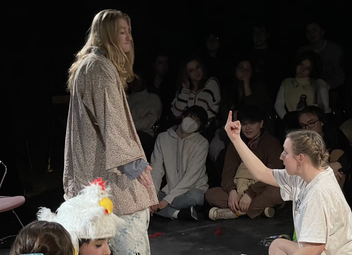 A young woman with blonde hair in braids is seated, raising her hand with one finger pointing up. A girl with long hair, standing in front of her, appears to be speaking or answering. Several people are watching in the background, some sitting on the floor, one wearing a chicken hat, and others standing.