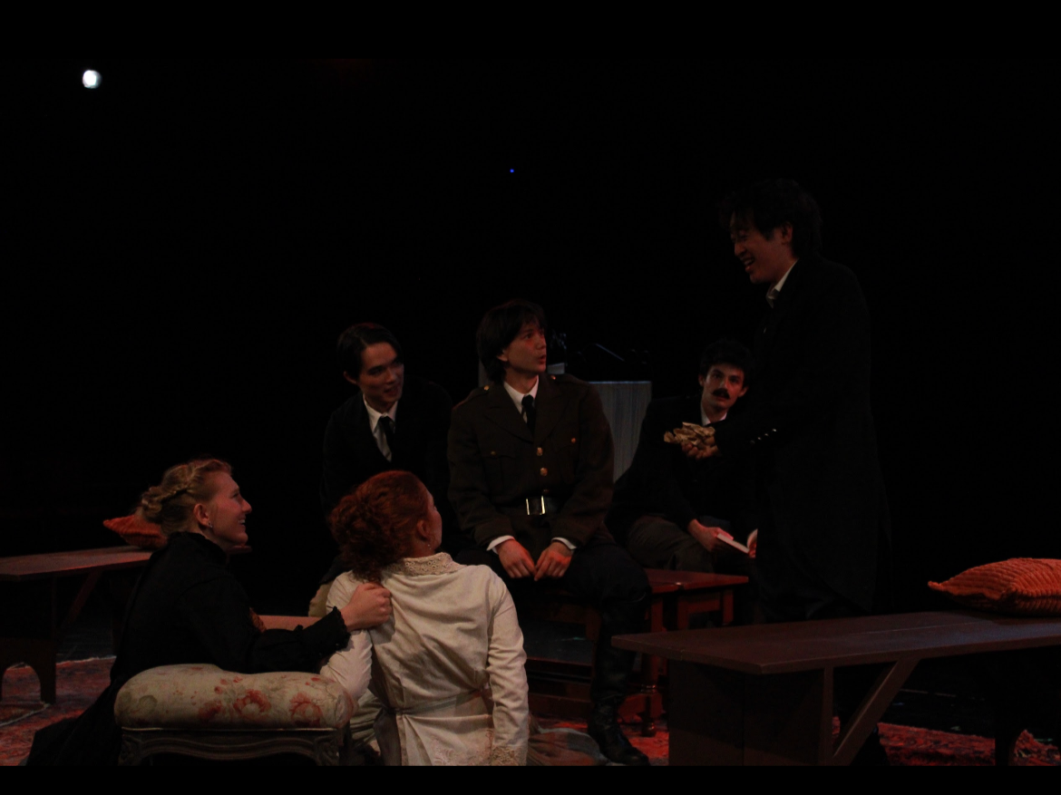 People in costumes performing in a theater, with some seated and one standing, engaging in a scene on stage.