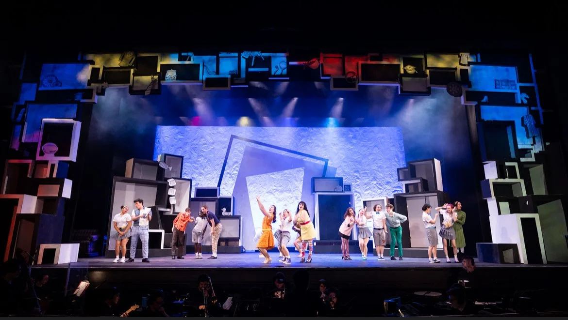 Theater stage with a group of performers dancing and singing, set with colorful geometric shapes and props.