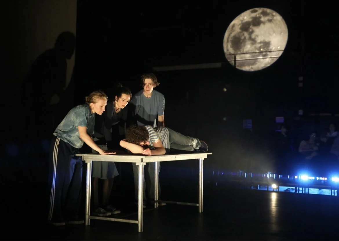 Four young people perform on stage, with one lying face down on a table and the others leaning over or standing around, under a projected image of the full moon in a dark setting.