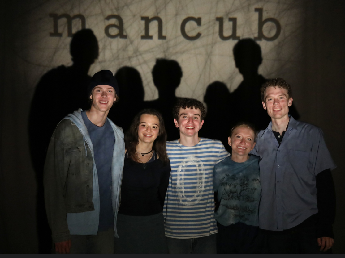 Group of five smiling young people standing in front of a wall with the word 'mancub' projected on it, casting shadows behind them.