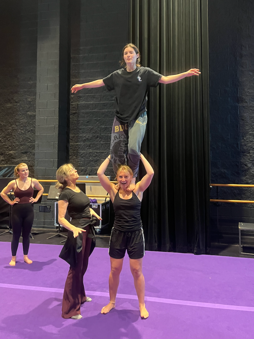 A group of women practicing acrobatics on a purple gym mat in a gym studio. One woman is standing on the shoulders of another, balancing with arms outstretched. Two other women stand nearby, watching and smiling.
