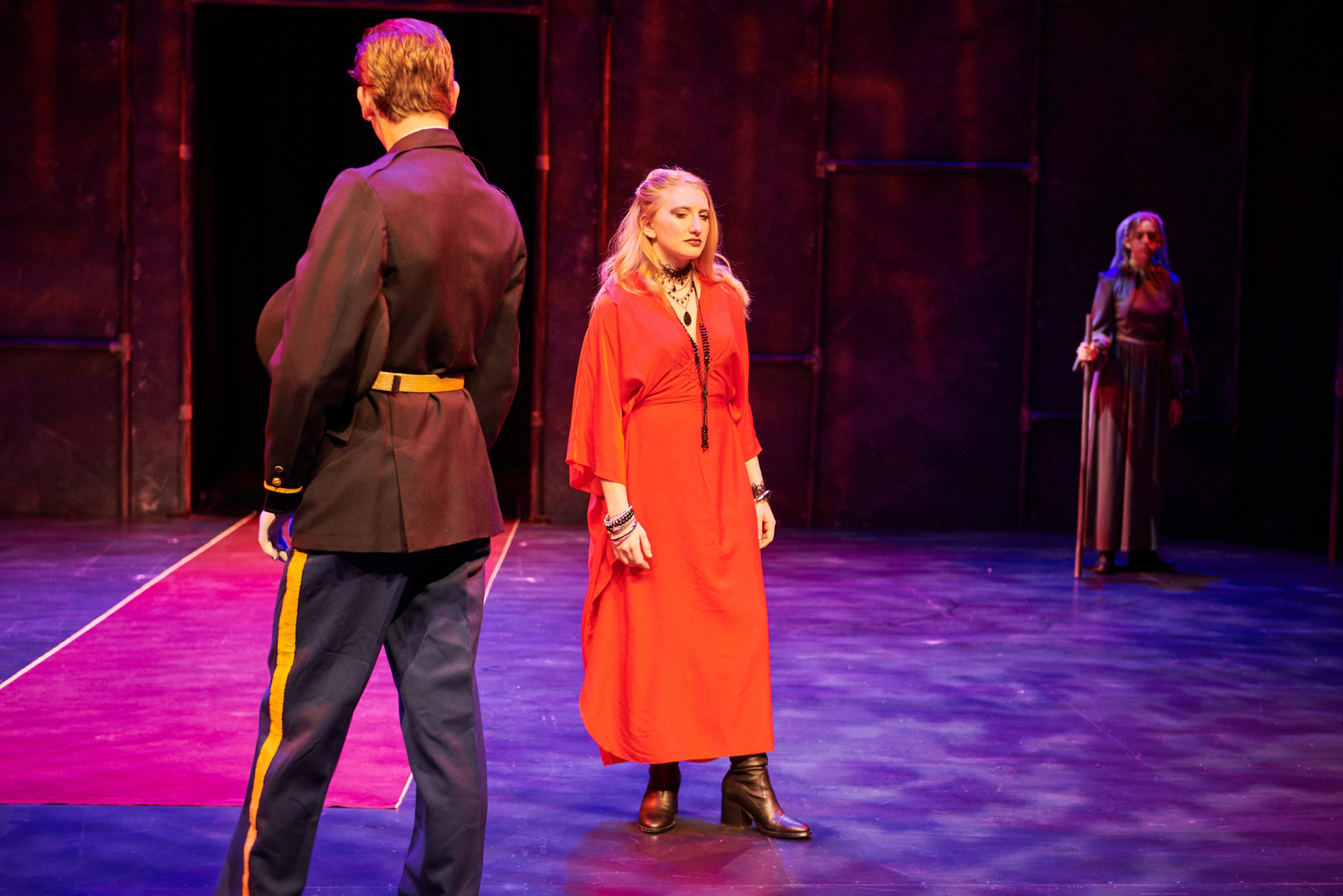 Three women on stage during a theatrical performance with colorful lighting. One woman faces away, dressed in a black uniform with a yellow stripe, another in a red dress with jewelry, and the third in the background in a long dark dress with a staff.