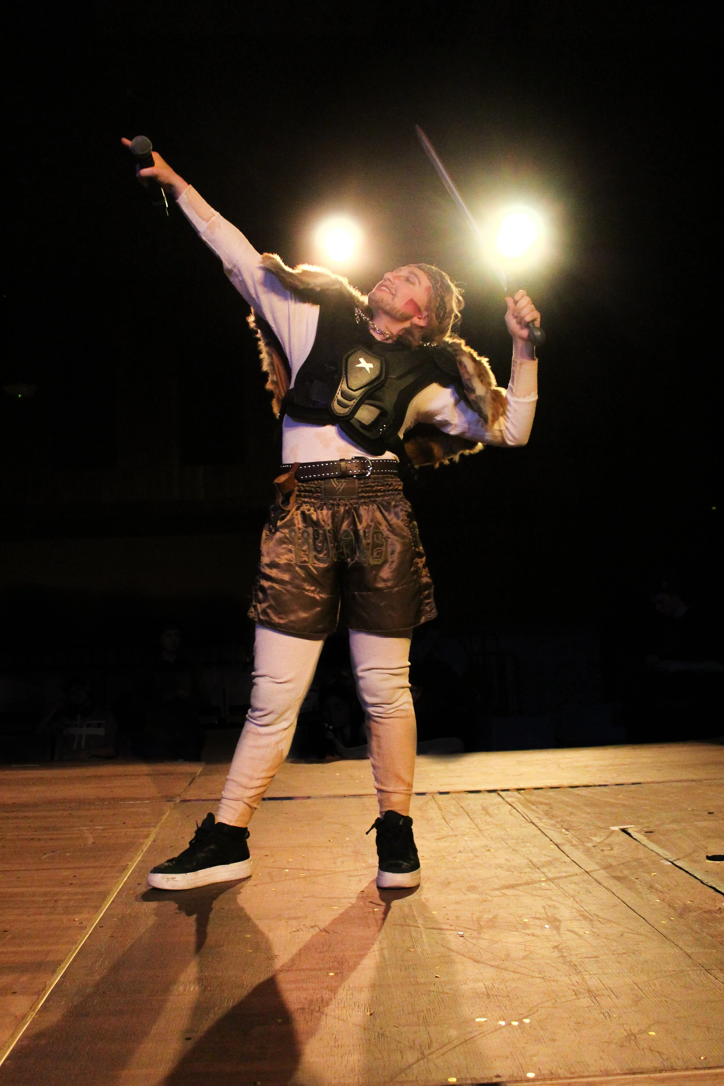 Performer in flashy clothing on stage, holding a microphone in one hand and a sword in the other, with bright stage lights behind, in a theatrical or entertainment setting.