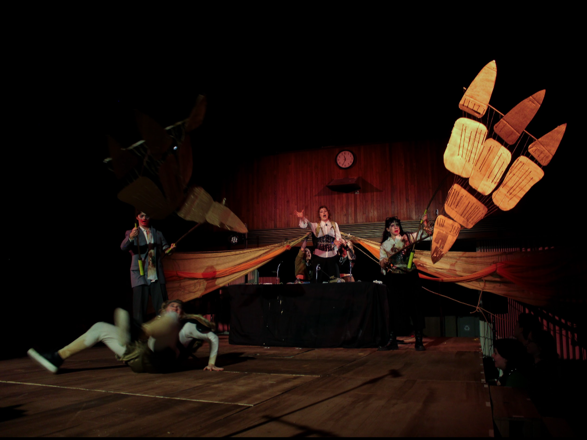 Theater performance with performers manipulating large paper or cardboard hand props, a woman singing at a microphone, and a person falling on the stage.