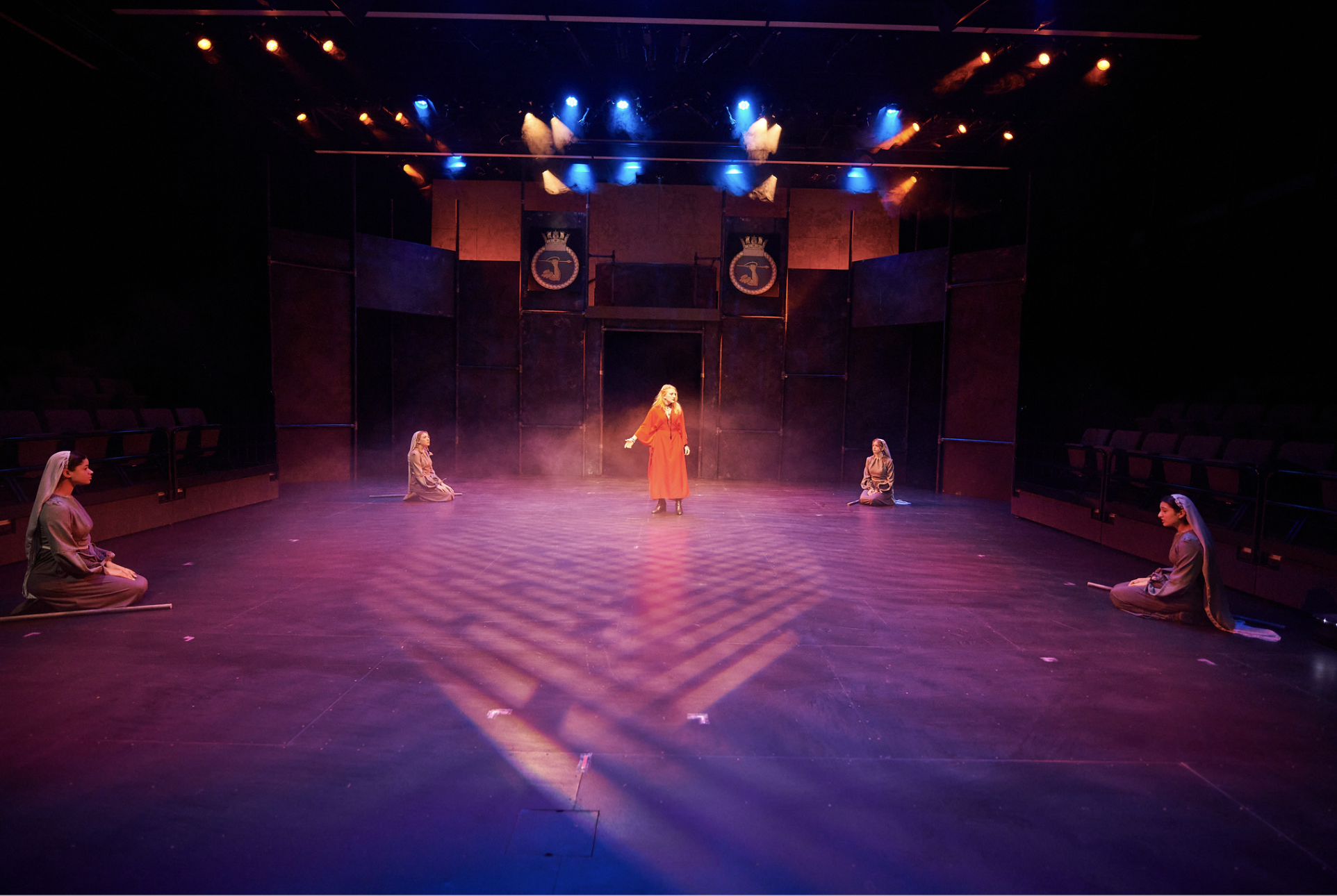 The stage of a theatrical play with five actresses in costume, one in the center wearing a long red coat and the rest kneeling on the floor in gray outfits with white veils. Stage lights cast purple, orange, and blue hues onto the scene, with two banners featuring crests hanging at the back.
