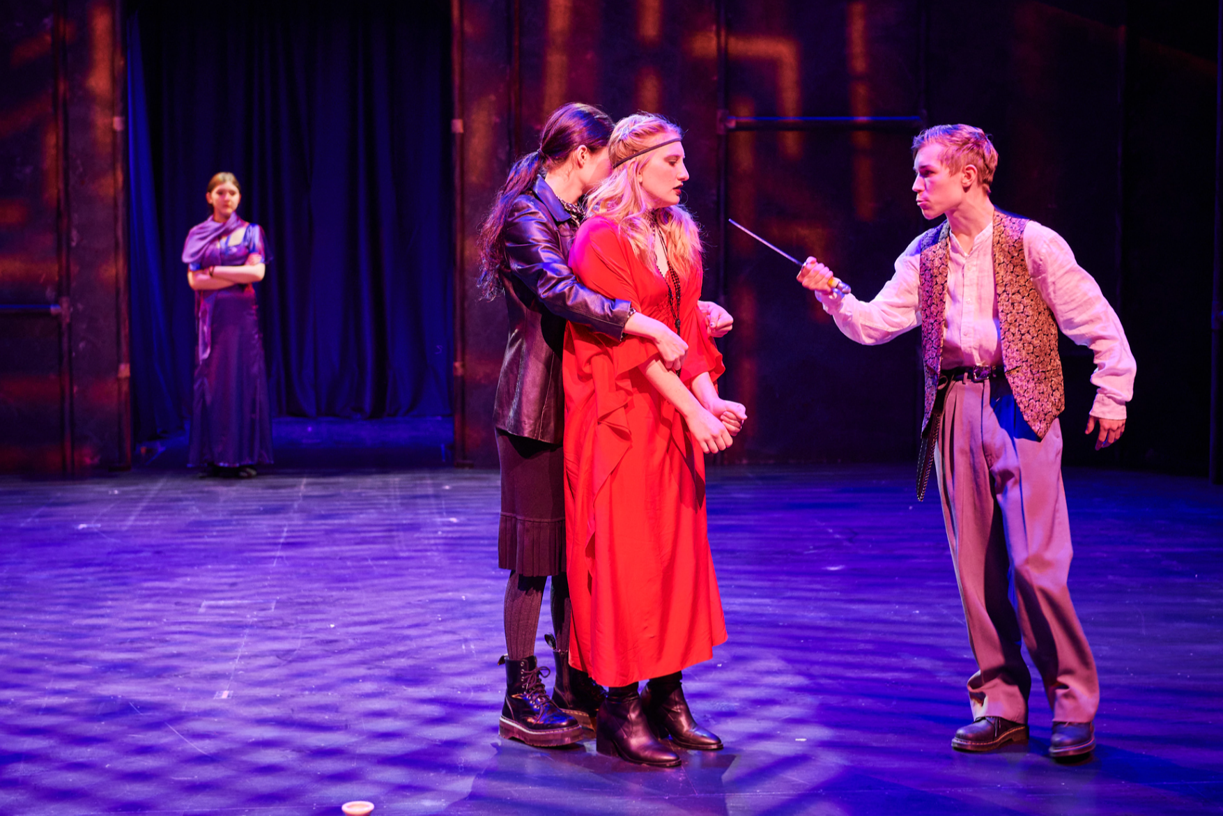 A theatrical scene with four women on stage. Three women are in the foreground, while one woman stands in the background. The woman on the right holds a sword pointing toward the woman in the middle, who wears a red dress. The woman on the left stands close to the woman in red, appearing to support her. The woman in the background is watching with her arms crossed, dressed in a purple outfit. The stage has dark curtains and wooden floor with purple lighting.