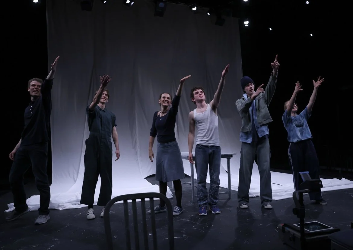 Group of six actors performing on stage with a white backdrop, some raising their arms, during a theatrical rehearsal or performance.