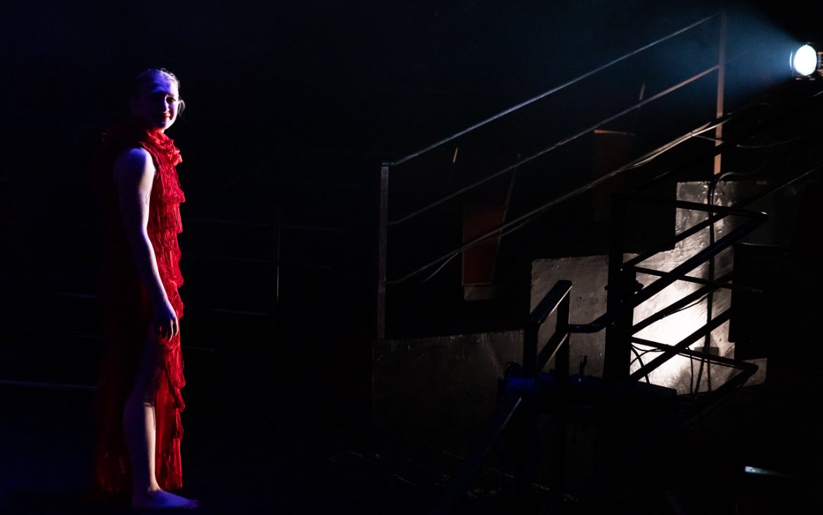 A woman in a red dress standing on a dimly lit stage with a spotlight behind her.