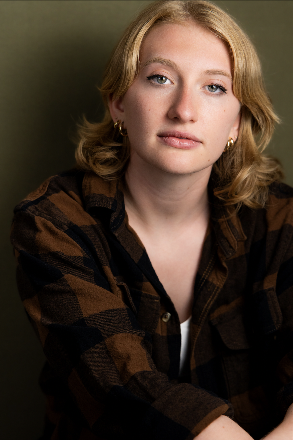 A young woman with blond, shoulder-length hair and fair skin. She is wearing hoop earrings and a brown and black checkered shirt, and has a neutral expression on her face, looking directly at the camera.