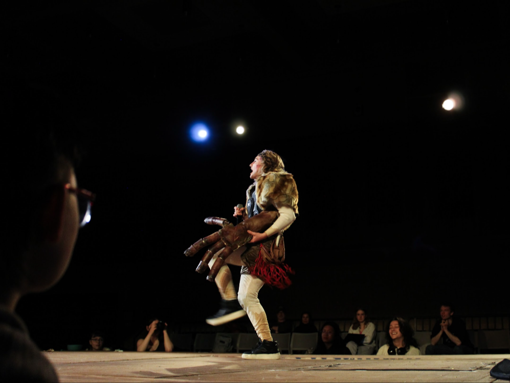 Performer on stage wearing a costume with a fur collar, holding a prop that looks like multiple tentacles, audience watching and smiling.