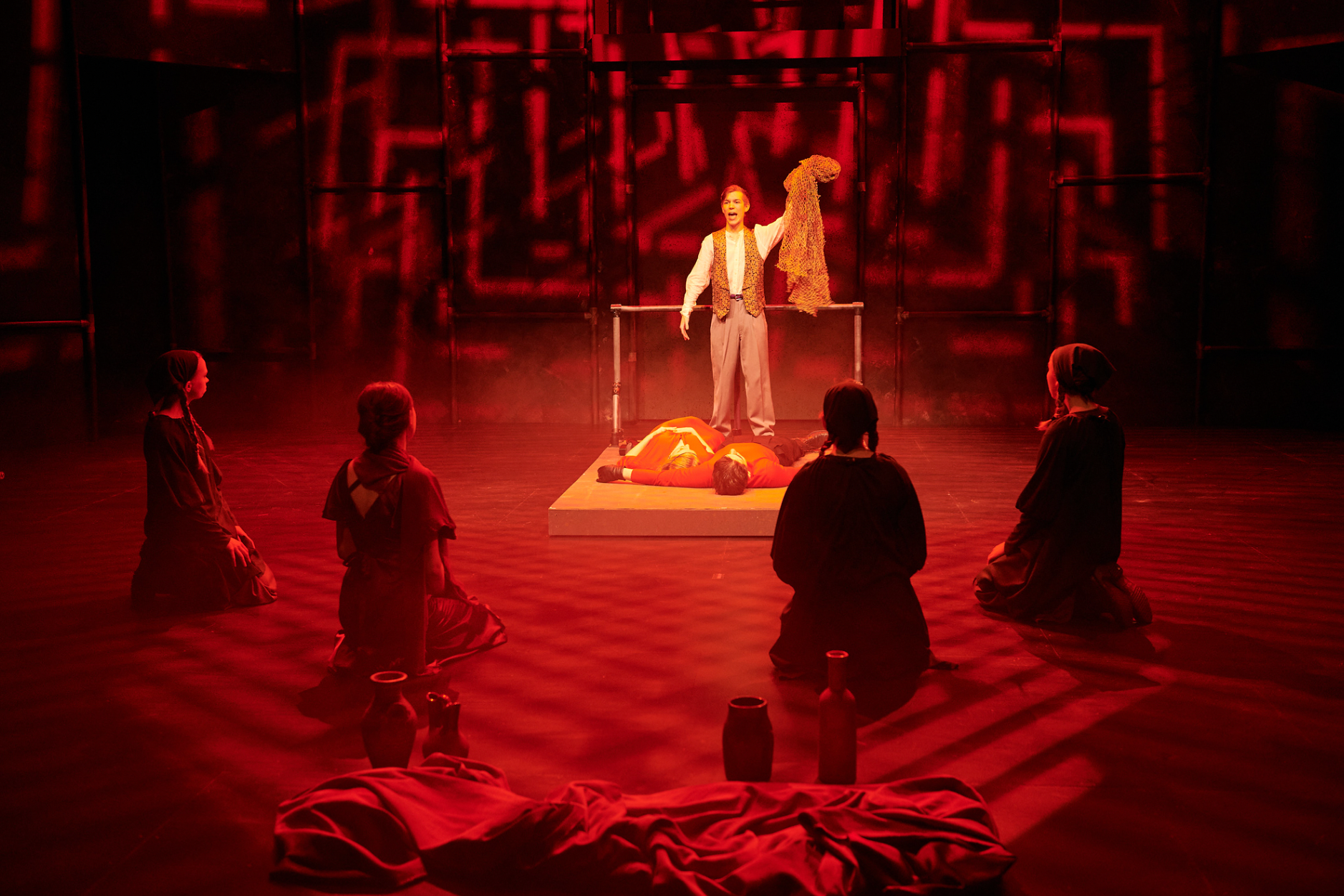 A theatrical performance with a woman standing on a platform holding a fabric, surrounded by four kneeling women and one lying figure, all in a red-lit stage with abstract background.
