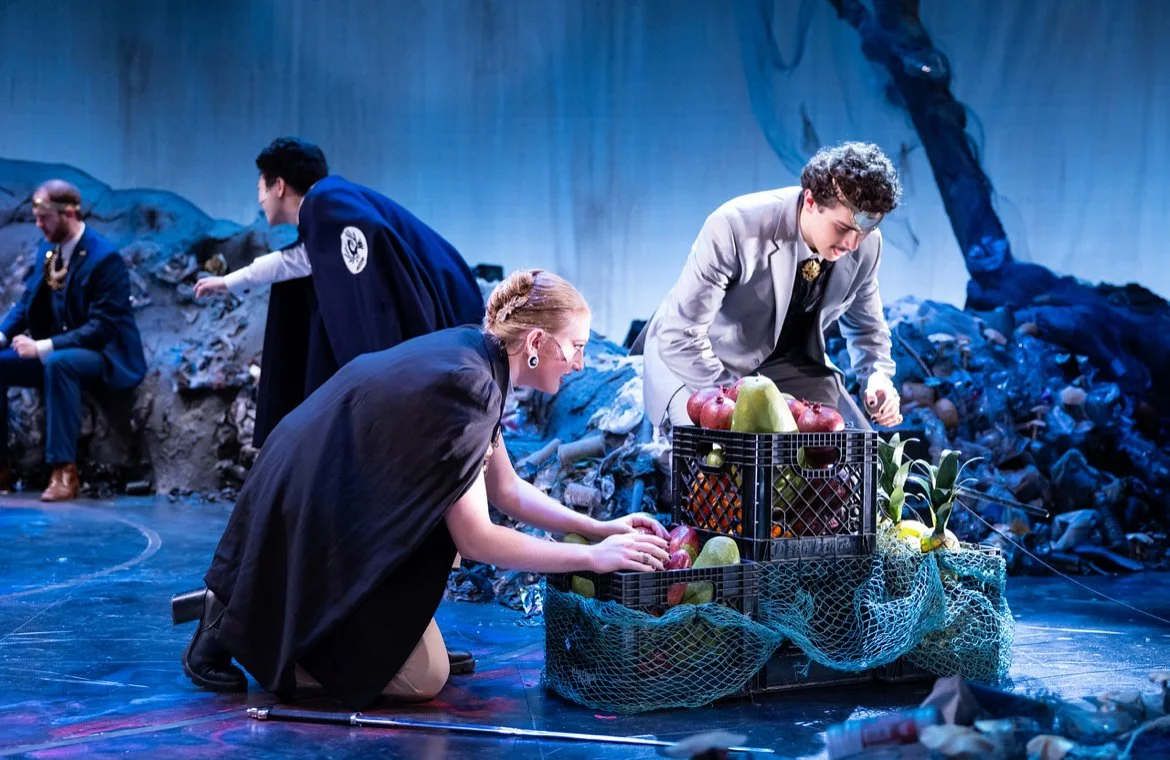 Four people on stage surrounded by trash and debris, with two of them examining a crate of fruit, including apples, pears, and pineapples.