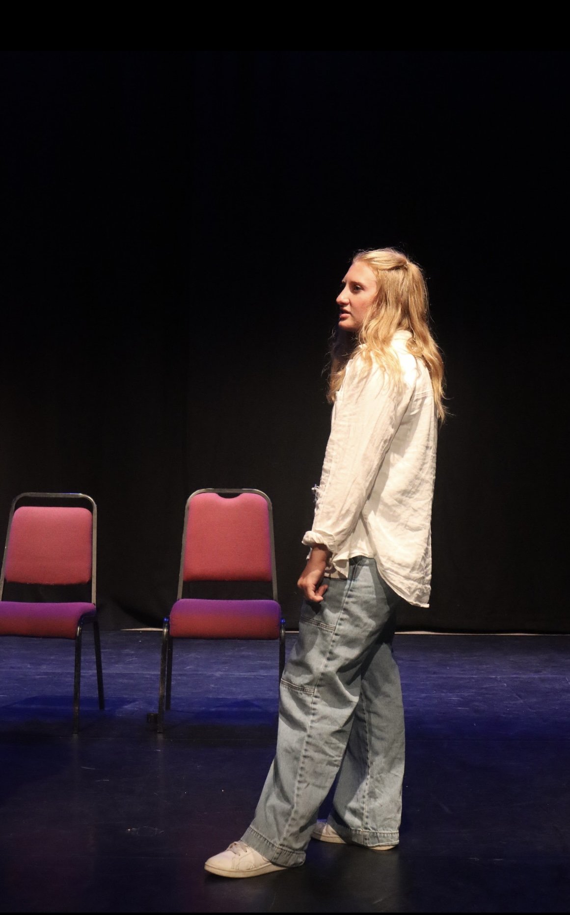 A young woman with long blonde hair, wearing a loose white shirt and baggy jeans, stands on a stage with a black background. Two red chairs with black metal frames are behind her.