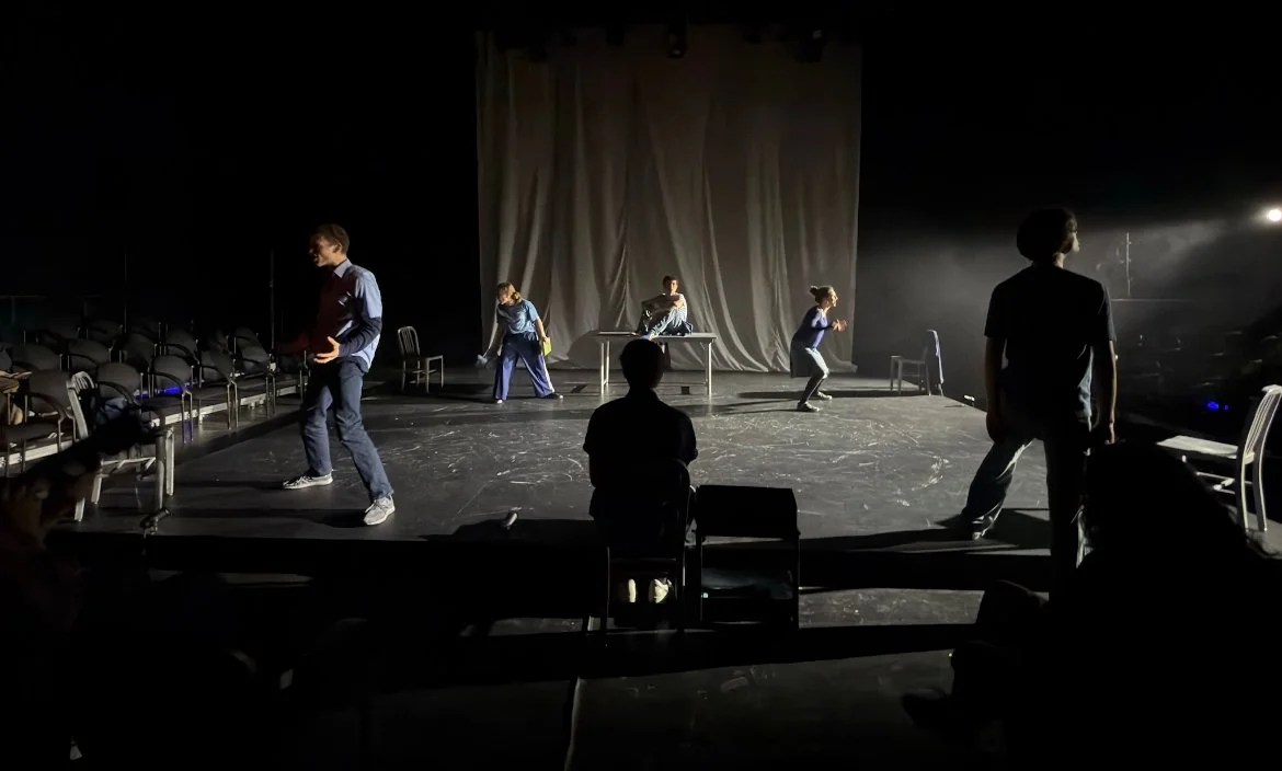 Theater stage with performers rehearsing, some standing and some seated, and empty chairs around the stage, with a backdrop curtain and dim lighting.