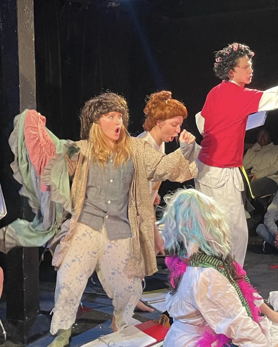 A group of actors performing on stage, wearing colorful costumes and wigs, with expressions of surprise or excitement, and an audience member visible in the background.