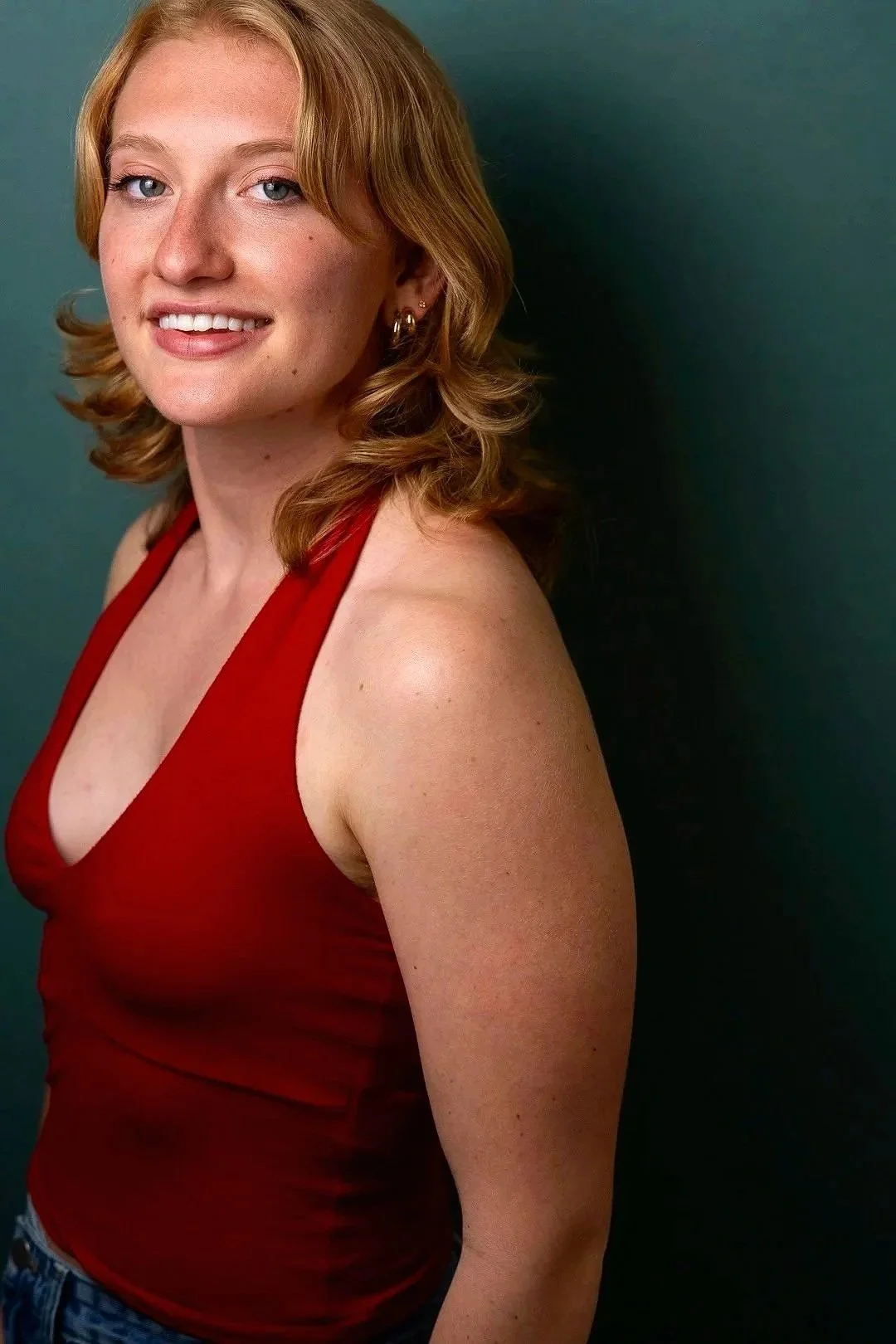 A young woman with red hair and light skin wearing a red sleeveless top, standing against a dark green background.