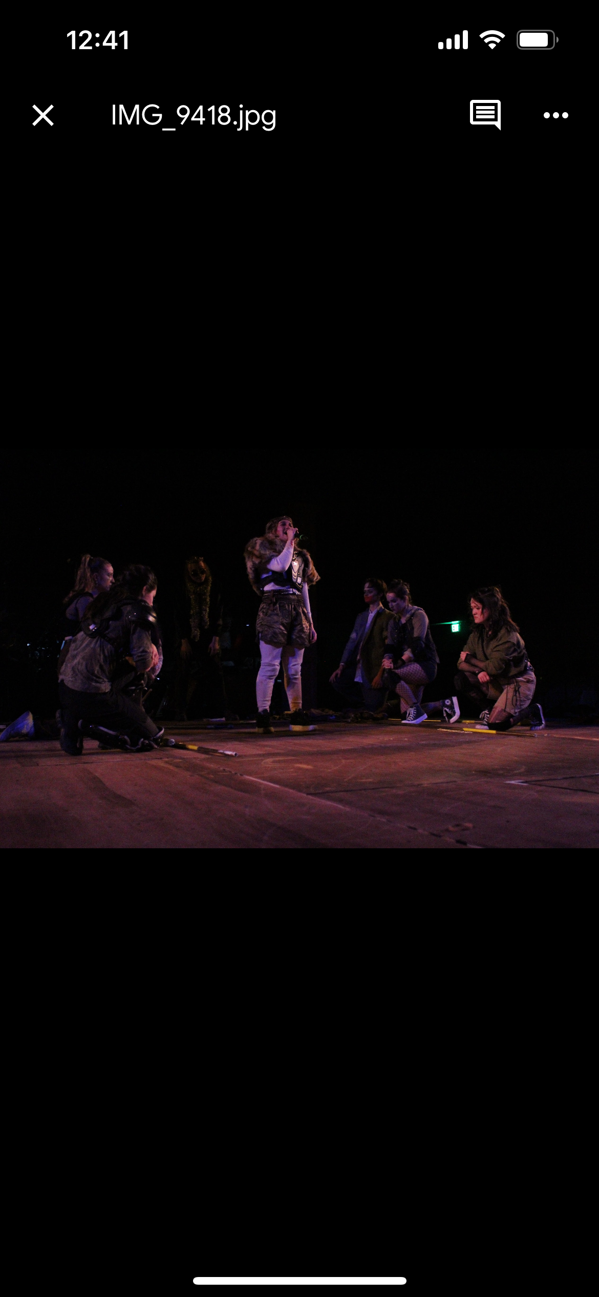 A woman with long hair, wearing a fur-lined jacket, standing on stage and singing into a microphone. Several women surrounding her are kneeling or squatting, looking at her, all dressed in casual or costume-like clothing in a dark auditorium with stage lighting.