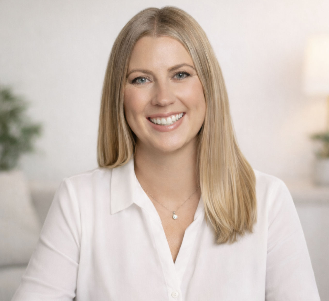A woman with long blonde hair smiling, wearing a white blouse and a silver necklace with a small charm, in a bright, modern indoor setting with plants in the background.