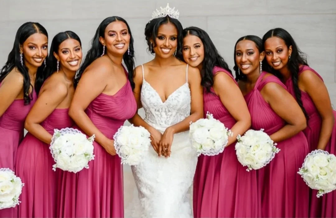 Bride in white wedding gown with tiara standing with six bridesmaids in matching magenta dresses, each holding a white bouquet, all smiling. #bridalmakeup
