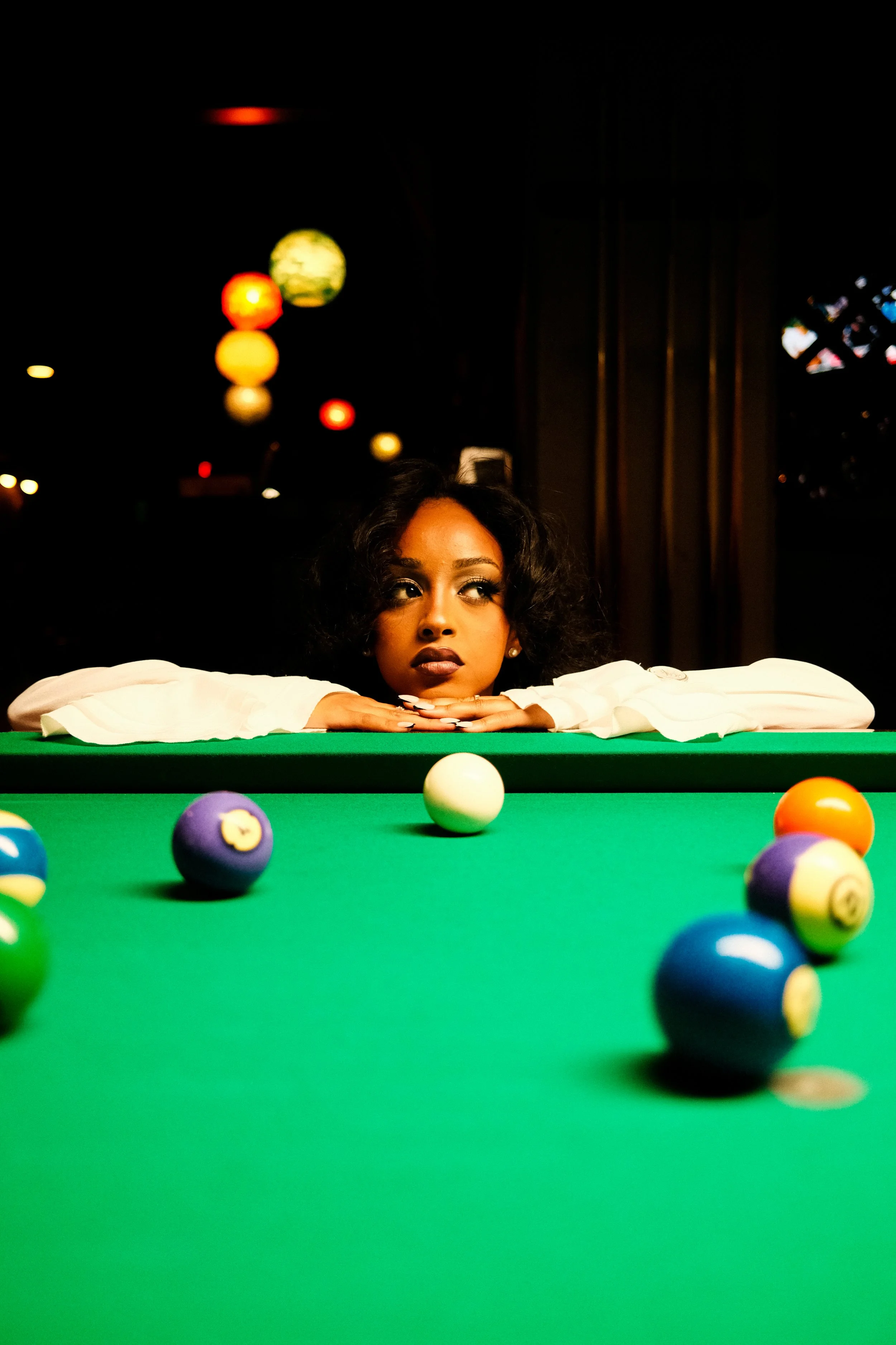 A woman rests her chin on her arms at a billiard table, looking contemplative, with pool balls scattered on the green felt surface, in a dimly lit room with hanging lights and dark curtains.