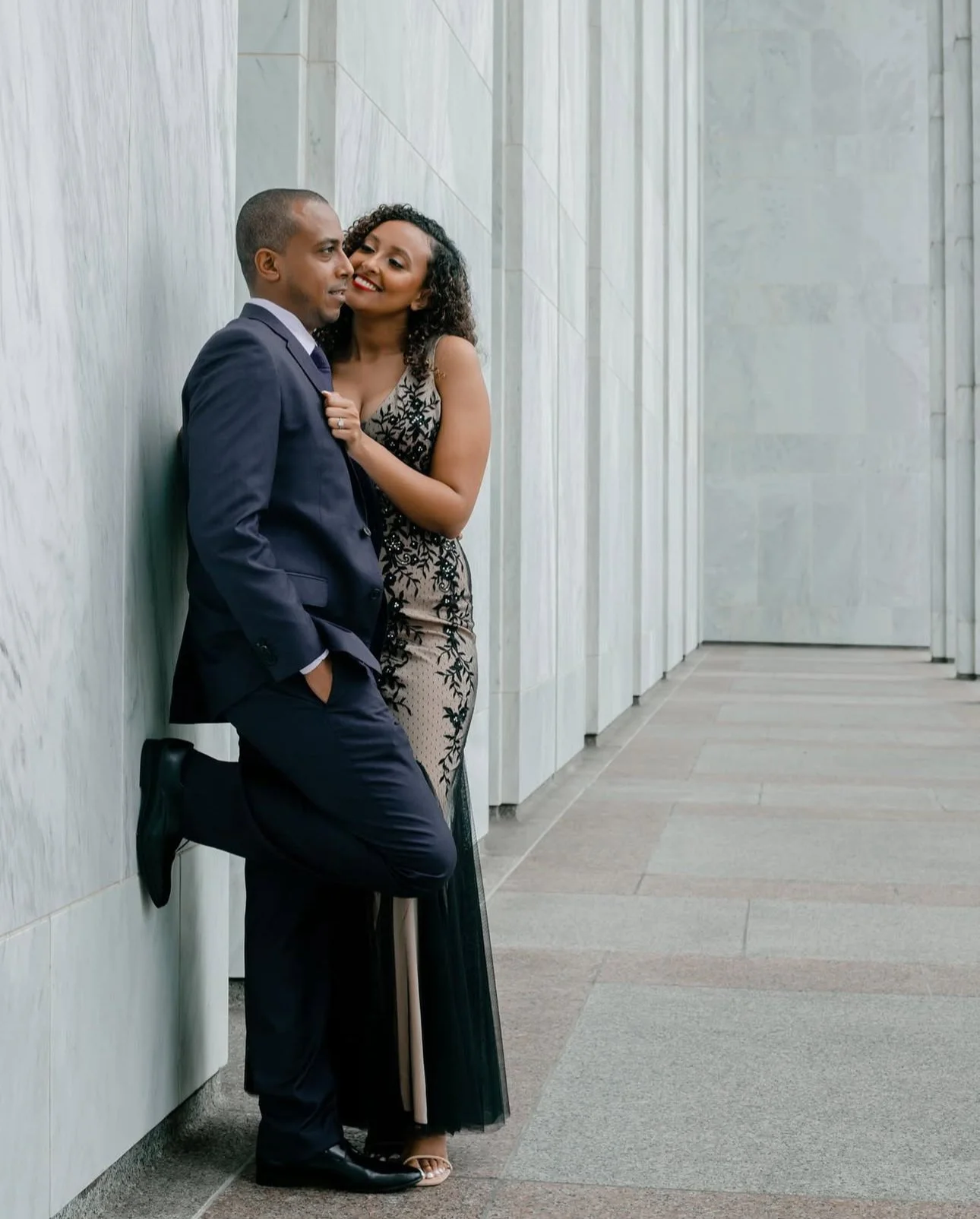 A man in a suit leaning against a wall with his hands in his pockets, and a woman in a black and beige dress with floral patterns, standing close and smiling at each other in a corridor with marble walls. #engagementphotos