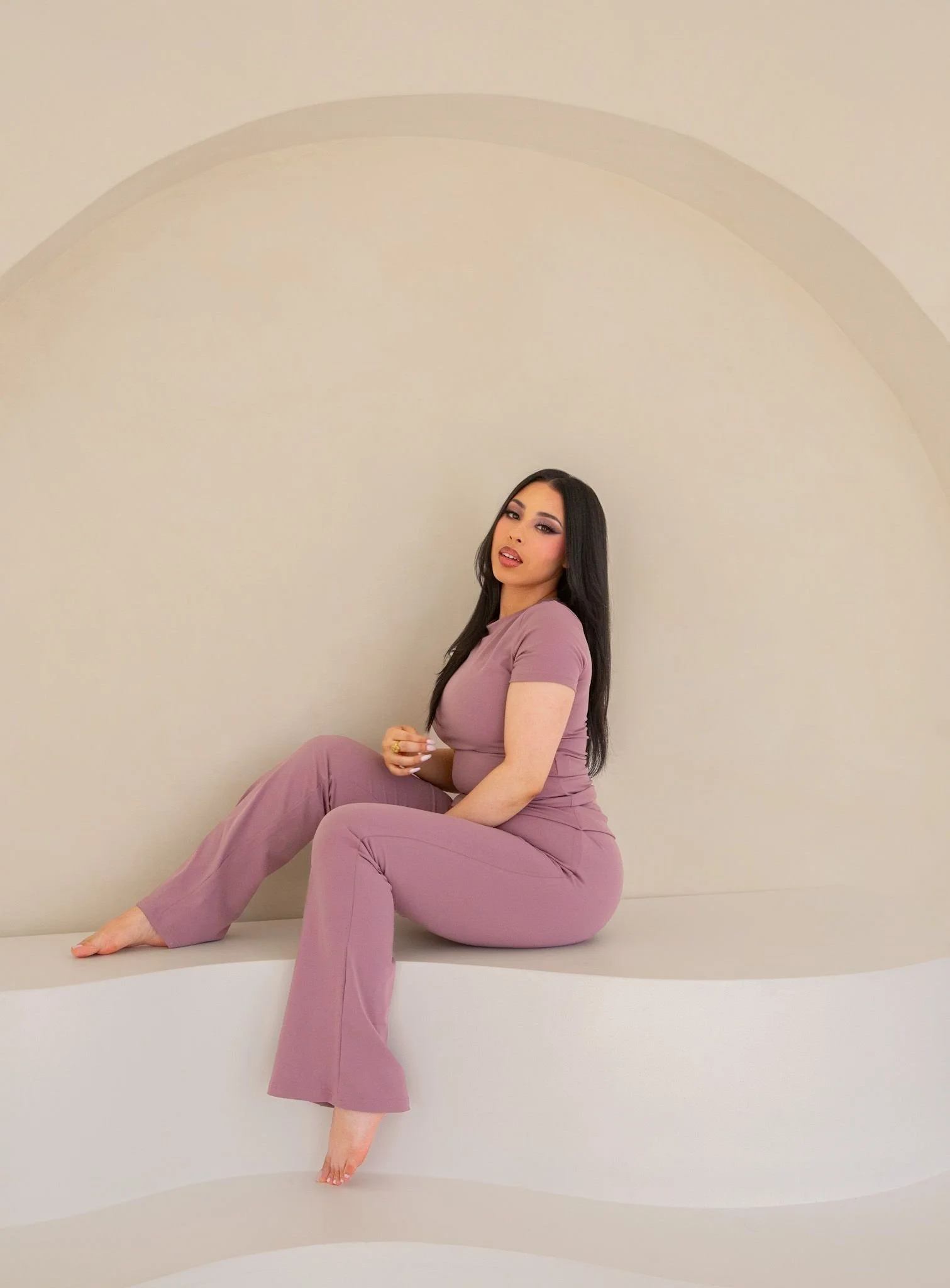 A woman with long dark hair wearing a mauve outfit sitting on a white curved surface against a beige wall with an arch design.