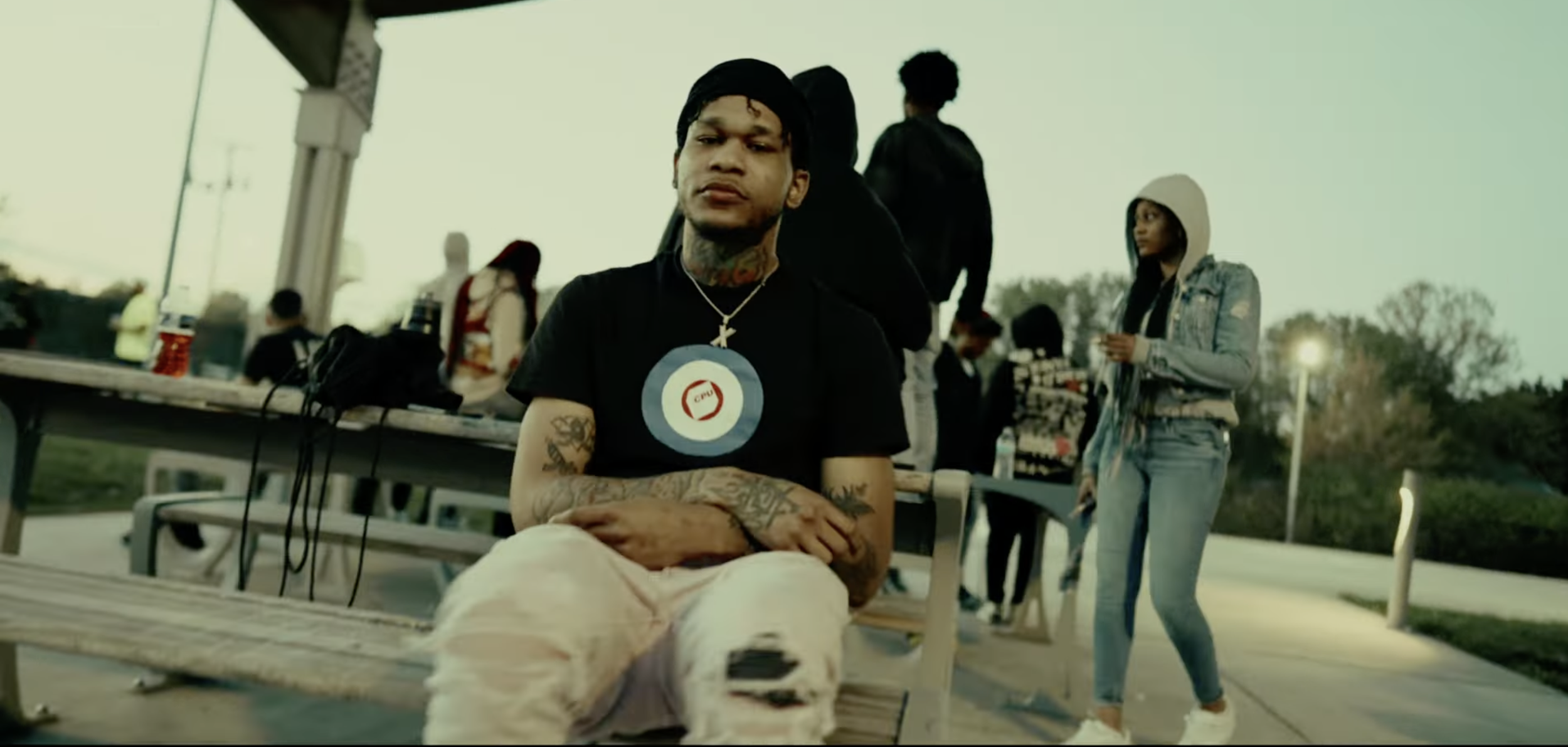 A young man with tattoos, wearing a black t-shirt with a red and white logo, and a black head covering, sits with his arms crossed on a park bench. Video by Kiirusly performed by Lil Xelly