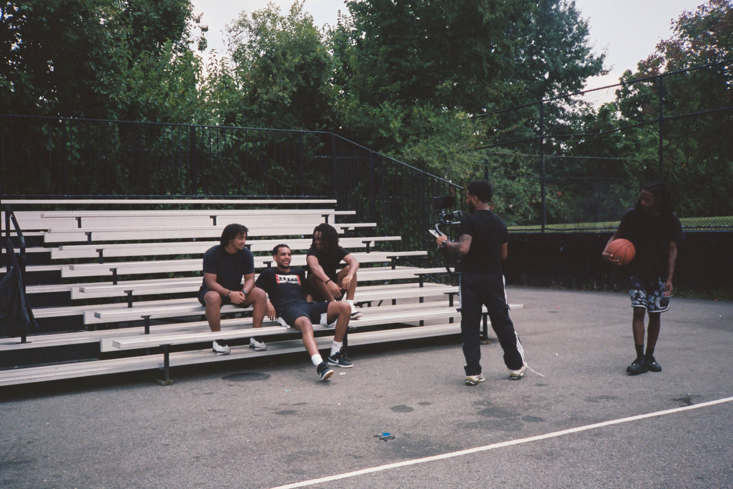 Four people sitting on bleachers at an outdoor basketball court, being filmed by a cameraman, with a person holding basketball nearby, surrounded by trees. Underground videographers