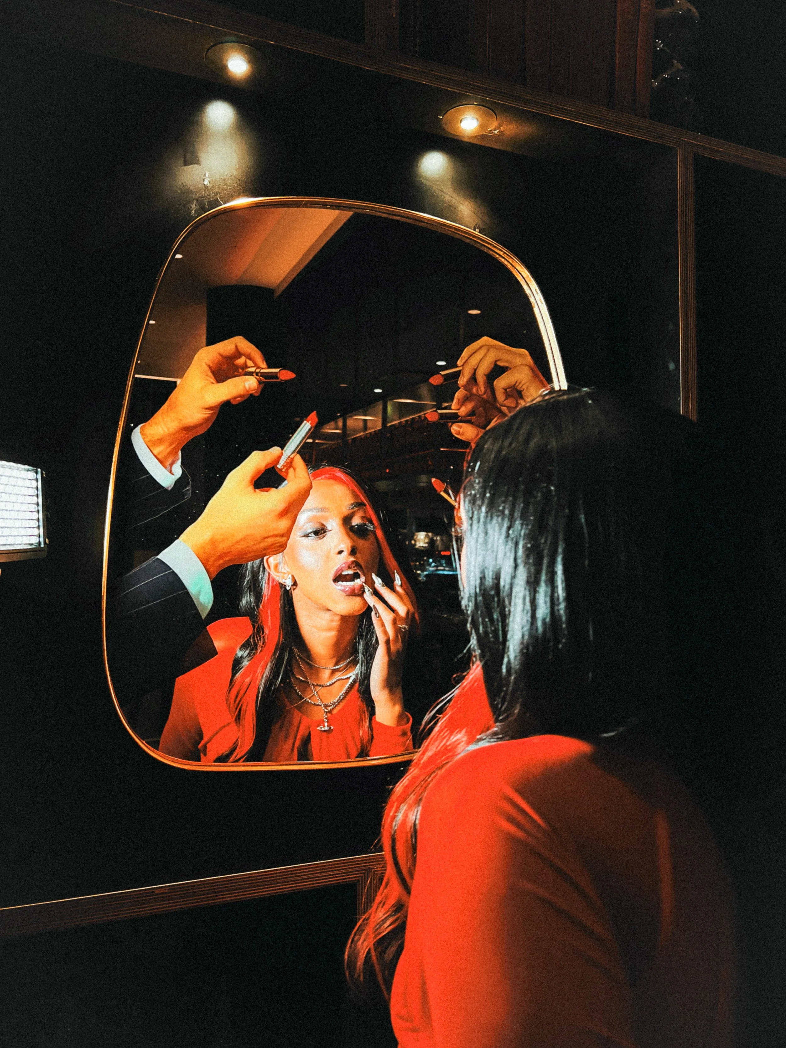 A woman with black hair sits in front of a mirror, having makeup applied by another person. The reflection shows the woman with bright red lipstick and layered jewelry, while the makeup artist is holding lipsticks and brushes.
