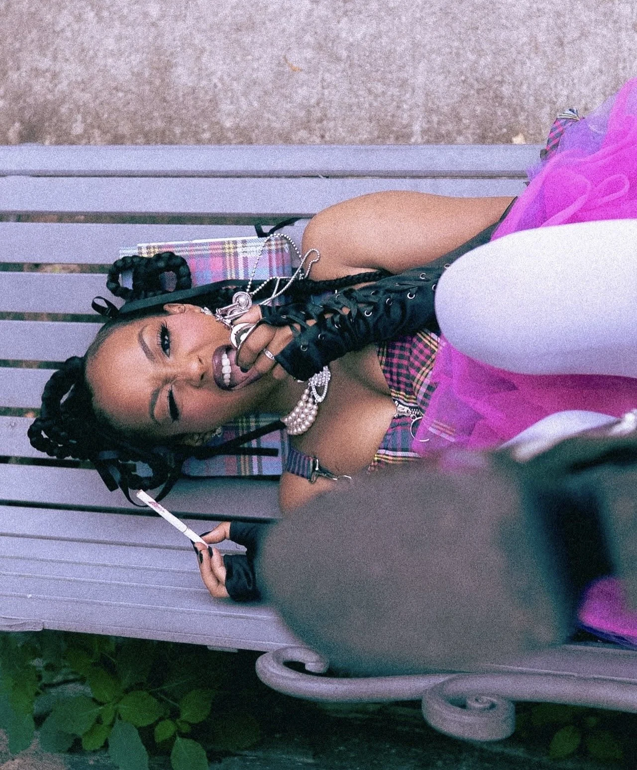 A woman with braided hair and makeup, wearing a plaid dress and jewelry, lying on a bench, covering her mouth with her gloved hand, with a pink and white outfit around her, looking at the camera.