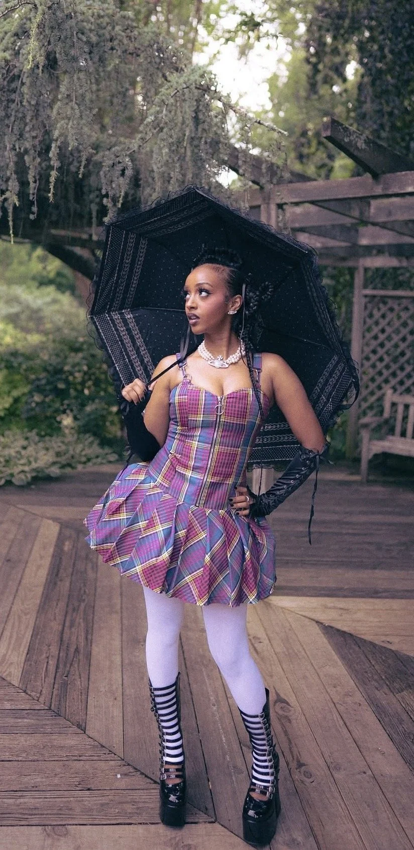 A woman with styled braids holding a black umbrella, wearing a colorful plaid dress with a ruffled skirt, white tights, striped high heels, black gloves, and jewelry, standing outdoors in a garden setting.