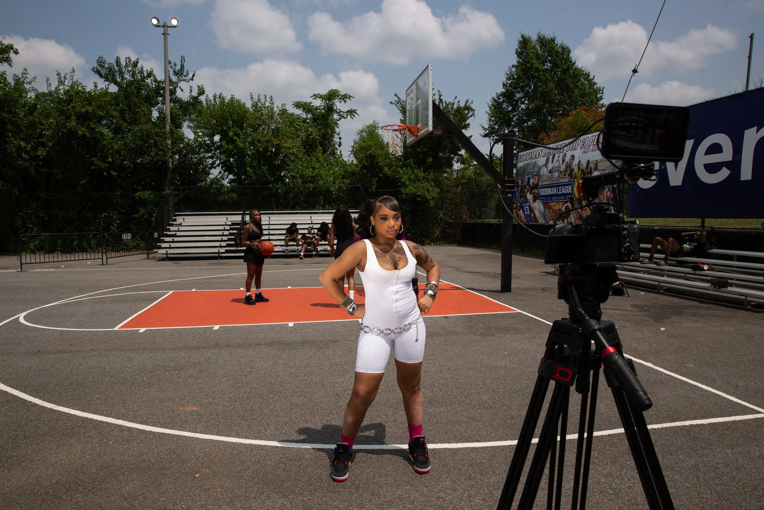 A woman in a white athletic outfit poses for a photo on an outdoor basketball court, with a camera and crew capturing the shot. Several women are in the background, some holding basketballs, and others seated on the bleachers.