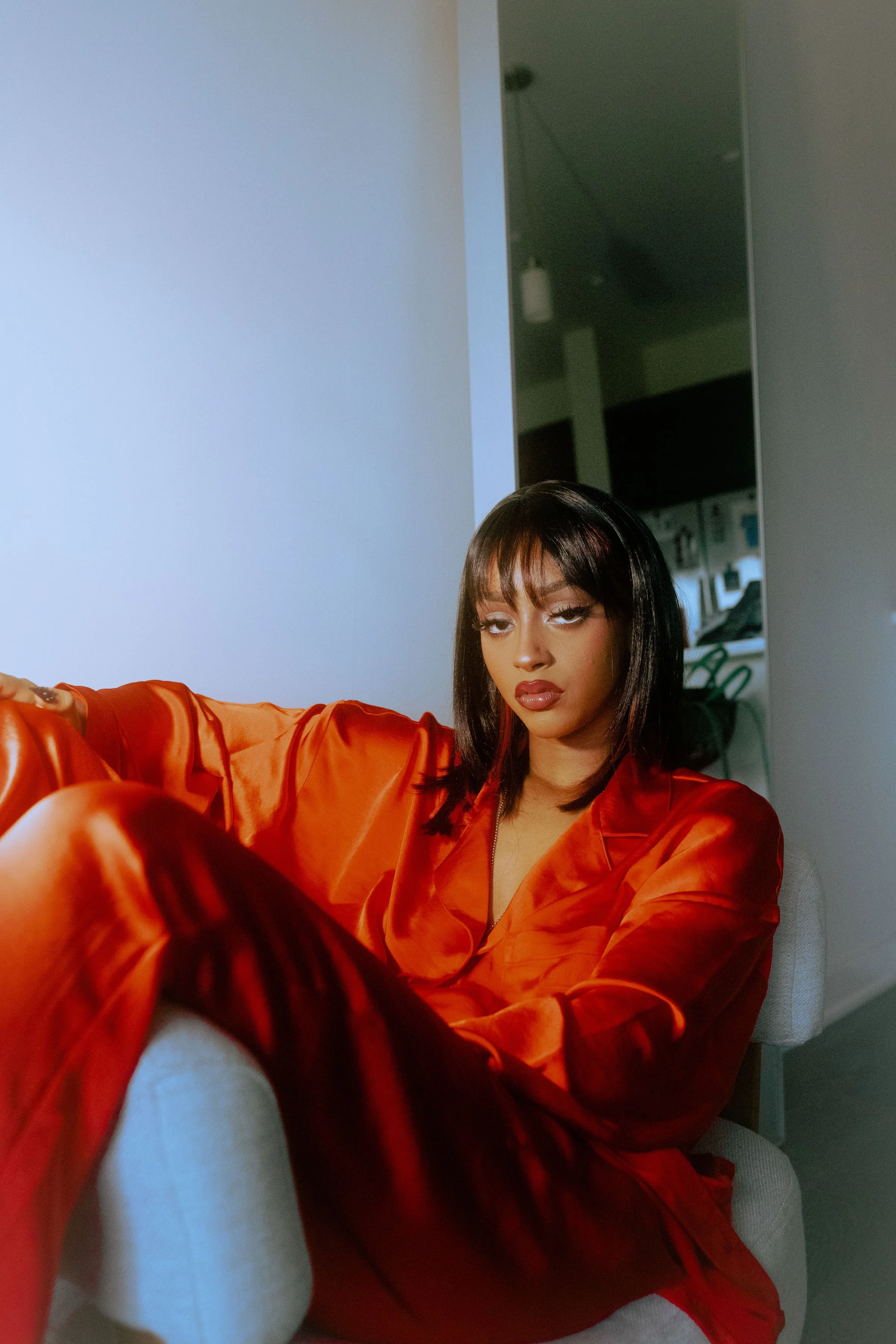 A woman with dark hair and makeup, wearing a satin orange robe, sitting on a light-colored armchair in a room with modern decor.