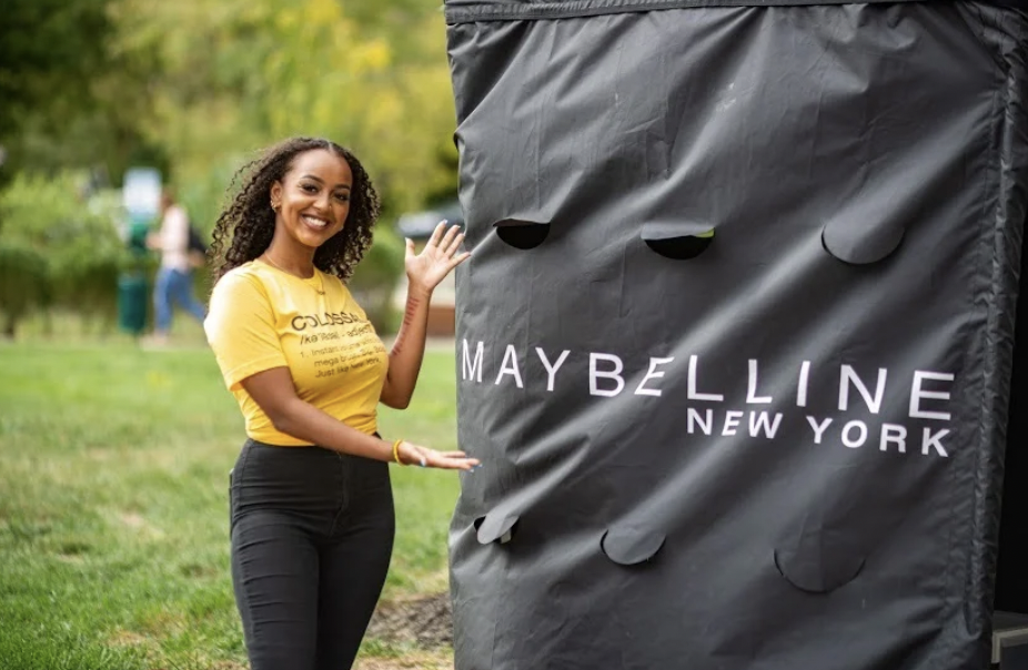Woman smiling and waving next to a large black Maybelline New York display or booth outdoors in a park. #MAYBELLINENEWYORK #MAYBELLINECOSMETICS @maybelline