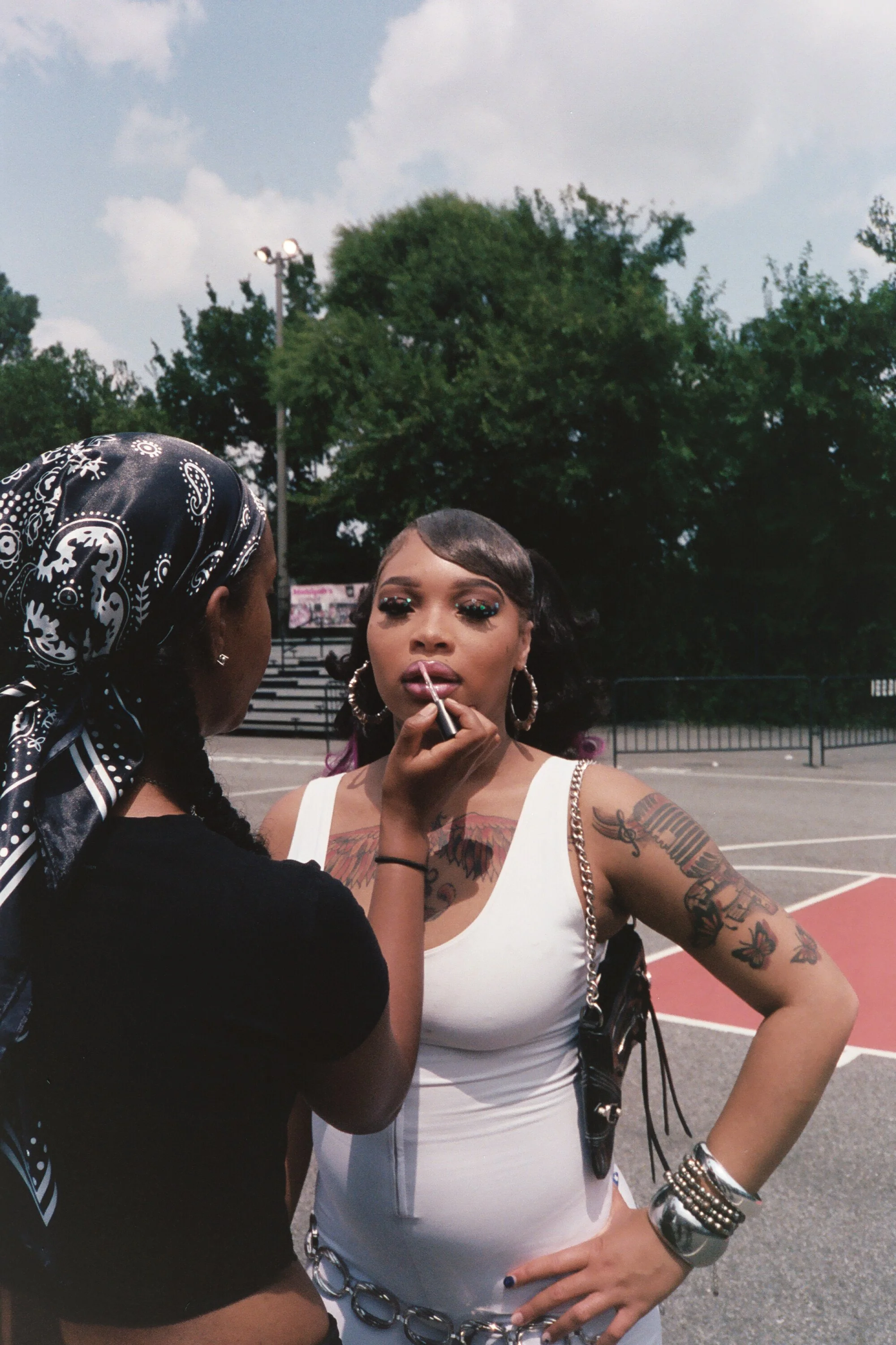 A woman with tattoos on her arm wearing a white tank top has her makeup applied by another woman with a bandana on her head, at an outdoor basketball court with trees and blue sky in the background.
