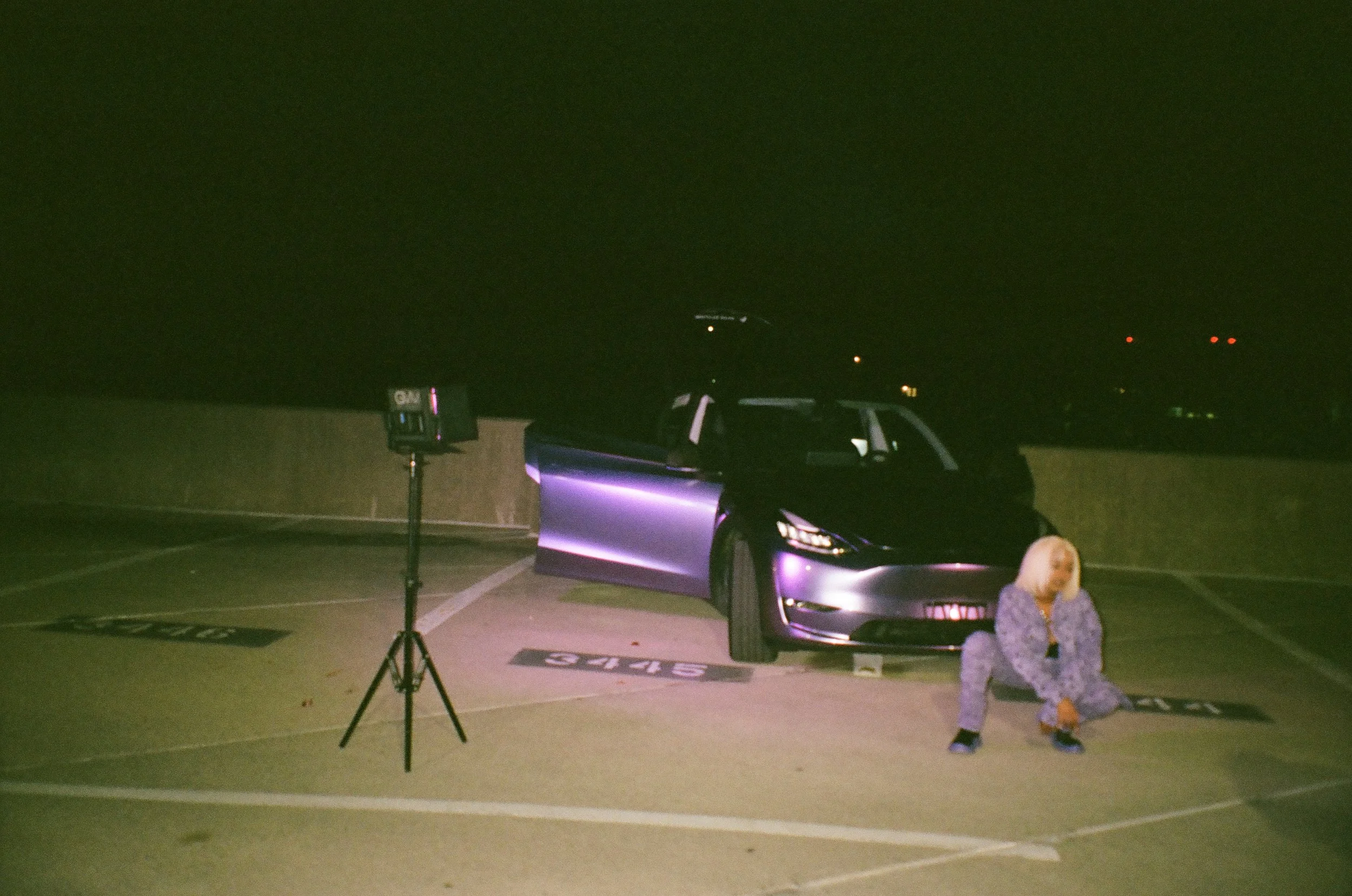 A woman with blonde hair, wearing pajamas, crouching in a parking lot next to a silver electric vehicle with purple accents at night, with a CNN sign on a camera stand nearby. #FANTA #FÁNTA music video @kiirusly